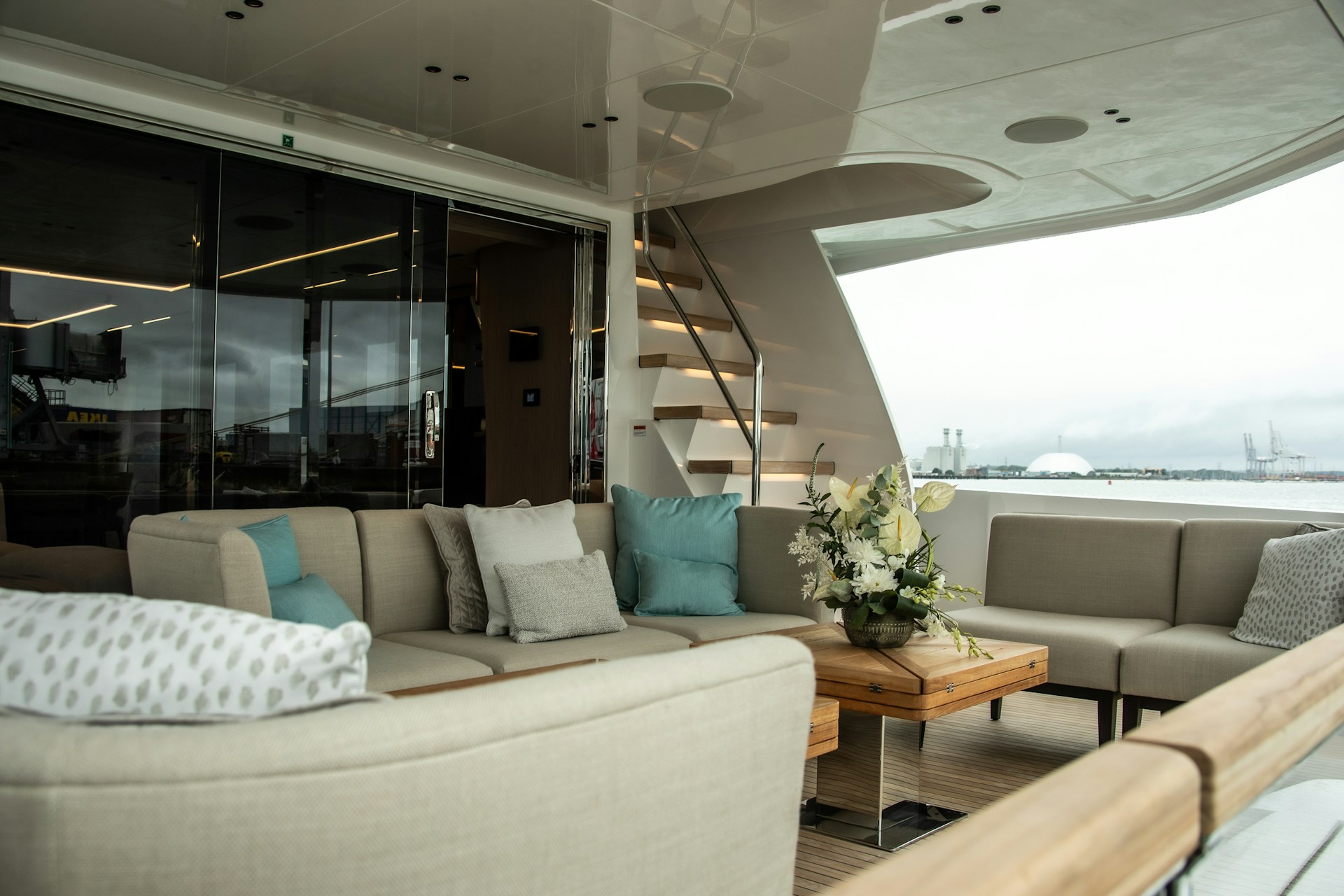 Luxury yacht aft-deck lounge with neutral modular seating, teak table, and harbor view—modern, sea-ready interior.