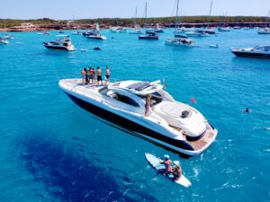A Mediterranean motor yacht at anchor with guests on the bow and turquoise water surrounding it.