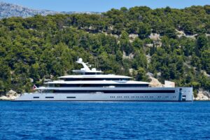 A modern white superyacht anchored off a pine-covered Mediterranean coastline, photographed by Arno Senoner.
