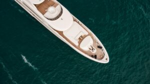 Aerial view of a superyacht gliding over deep blue water with guests relaxing on teak decks, symbolizing refined etiquette and quiet luxury.